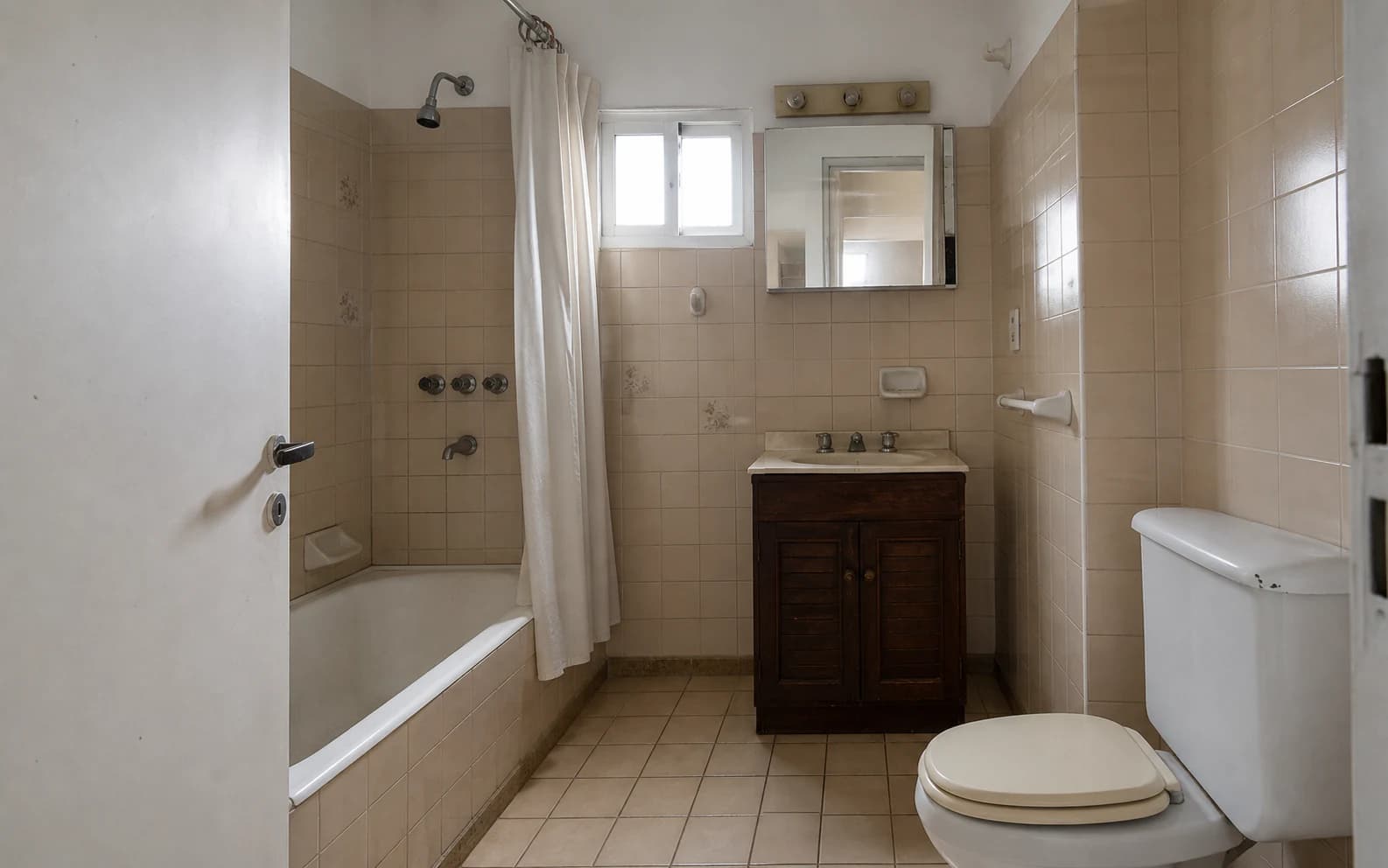 Mismo baño antes de la remodelación, con azulejos beige, bañera con cortina y mobiliario antiguo